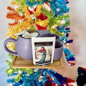 A bamboo tray with a purple tea set with a teapot and two cups and a bag of Sandia Spice tea on it in front of a rainbow colored Christmas tree with silver ornaments.