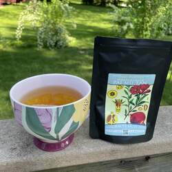 A bag of Pomegranate White Tea next to a teacup with brewed tea in it, on the edge of a railing with a beautiful green yard in the background.