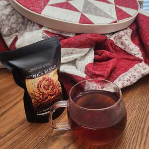 Monk's Blend black tea brewed in a clear glass mug with a bag of the loose leaf tea next to it and an in progress quilt in an embroidery hoop in the background.
