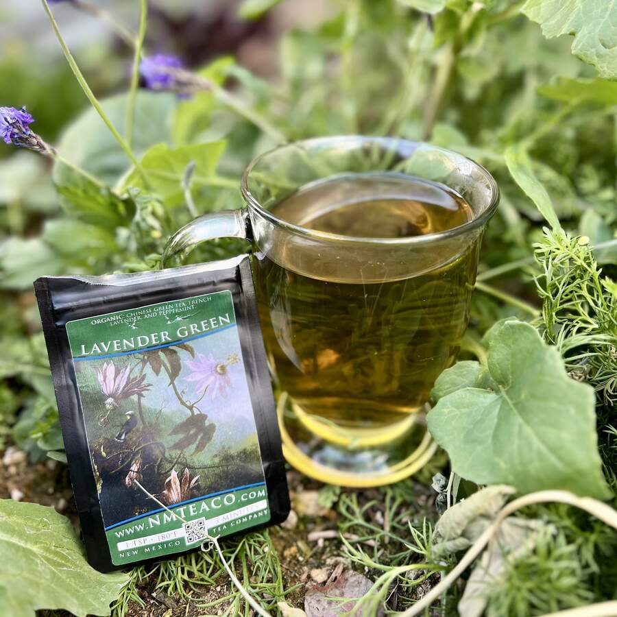 A bag of Lavender Green tea leaning against a clear glass mug full of brewed tea in a lush garden, surrounded by greenery.