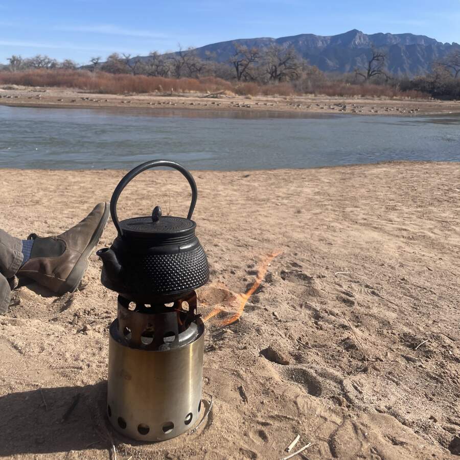 A black tetsubin iron teapot over top a makeshift stove with twigs burning below the pot with the Rio Grande and the Sandia Mountains in the background.