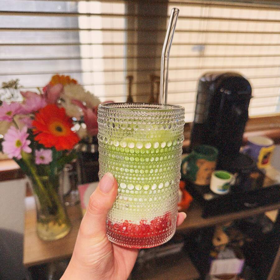 A hand holding up a glass of iced matcha with red syrup at the bottom of the cup and a glass straw in it.