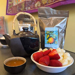 A bag of Grapefruit Pai Mu Tan white tea next to a black teapot with a bamboo handle behind a small cup of brewed tea and a bowl of strawberries and a kind of puffed cereal called sorghum.