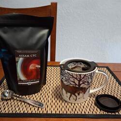 A bag of assam ctc tea next to a mug with an infuser brewing the tea and a 1 cup of perfect tea measuring spoon.
