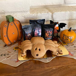A homemade spider-shaped bread loaf in front of bags of English Breakfast, Chocolate Black, and Hazelnut Black tea, with a few pumpkin and bat halloween decorations in the background.