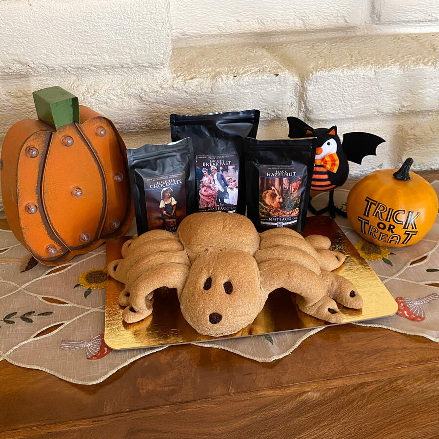 A homemade spider-shaped bread loaf in front of bags of English Breakfast, Chocolate Black, and Hazelnut Black tea, with a few pumpkin and bat halloween decorations in the background.