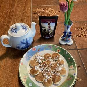 A plate of tiny, pancake-like poffertjes covered in powdered sugar with a teapot, a bag of Dutch Licorice Tea, and a vase of wooden tulips in the background.