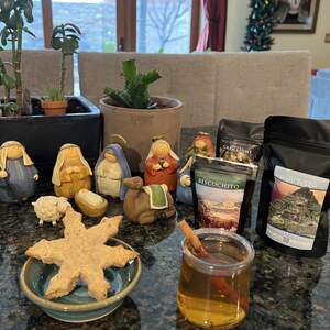  A brewed cup of biscochito tea with a whole cinnamon stick in it, and a snowflake shaped biscochito cookie beside it. In the background, there is a cute little nativity scene and three bags of tea including Biscochito green tea, machu peachu white tea, and monks blend black tea.