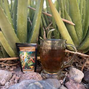 A clear glass mug full of brewed Baya Bonita Mate next to a bag of loose leaf Baya Bonita with a graphic label. In the background is an aloe plant.