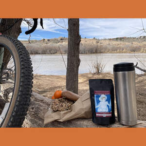 On the side of a river there is a bike parked next to a fallen log that has a couple of bagels, a couple of clementine oranges, a bag of Blueberry Bang Rooibos and a silver travel tea tumbler.