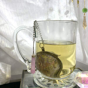 A clear glass mug with a tea ball infuser, with a rose quartz crystal point attached to the chain, full of Lavender Chamomile tea being steeped.