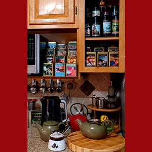 The corner of a kitchen with an open upper cabinet and a shelf full of loose leaf tea in tins with the graphic labels on them. Underneath the cabinet, on the countertop, is a collection of teapots, tea kettles, and infusers.