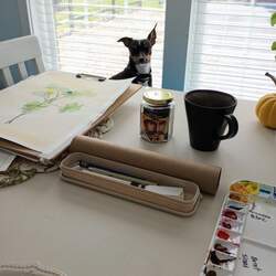 A small dog watching the photographer from the other side of a table covered in a beautiful painting and painting supplies with a jar of Cream Earl Grey tea and a mug of brewed tea in the center of it all.