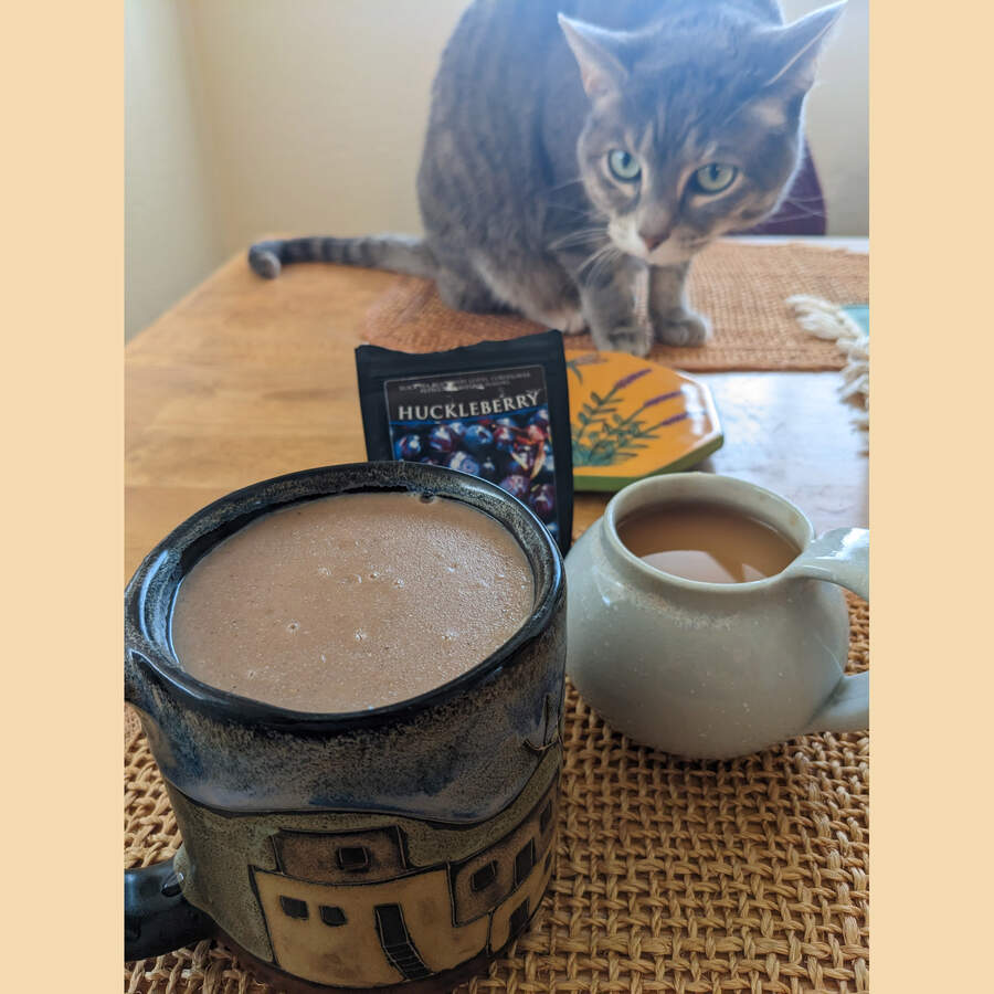 A cat in the background sniffing a brewed mug of Huckleberry Black tea with the bag of tea next to it and a mug full of atole for breakfast in the foreground.