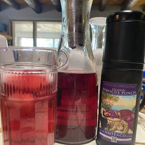 A glass and a jug full of Island Hibiscus Punch sun tea next to a tin of the loose leaf tea.