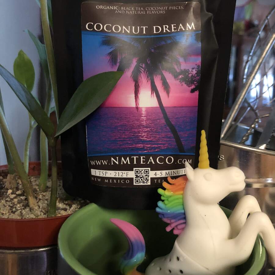 A bag of Coconut Dreams black tea with a mug that has a rainbow unicorn silicone infuser hanging over the side to steep the tea.