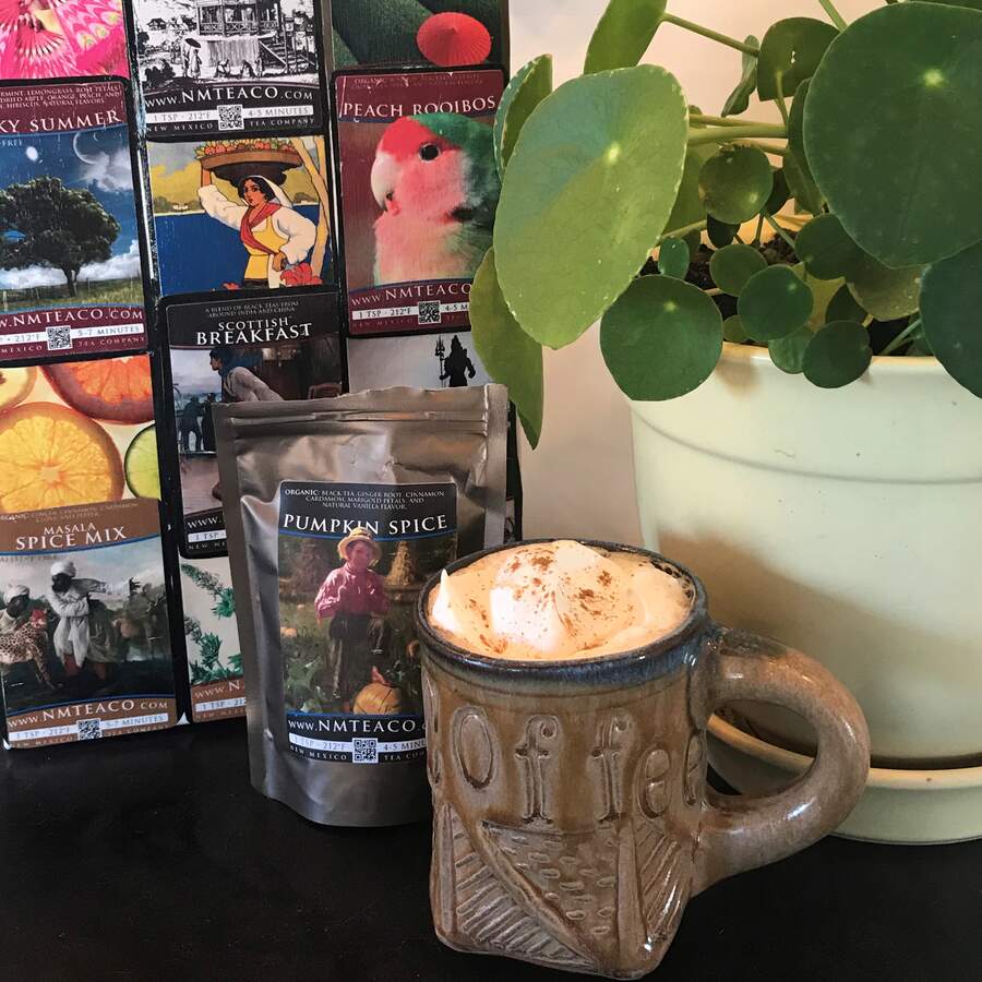 A brown, handmade mug filled with a Pumpkin Spice Black tea latte with whipped cream on top and a sprinkle of cinnamon. Behind it is a bag of Pumpkin Spice Black tea and a potted plant. In the further background is a display of various tea labels for other teas.