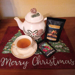 A festive teapot with pine needles, pinecones, and holly painted on it behind a teacup full of tea on a saucer, next to bags of loose leaf Maple Pecan Danish and Cranberry Black teas, all sitting on a Christmas placemat with Christmas trees and a script greeting of 