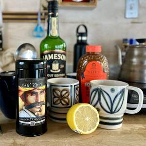 A tin of Cream Earl Grey tea next to two painted mugs, a bottle of Jameson, a bottle of honey, and half a lemon.