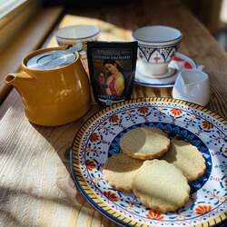 A photo of shortbread cookies on a plate with a bag of Golden Orchard tea behind it, next to a small yellow teapot with a couple tea cups in the background.