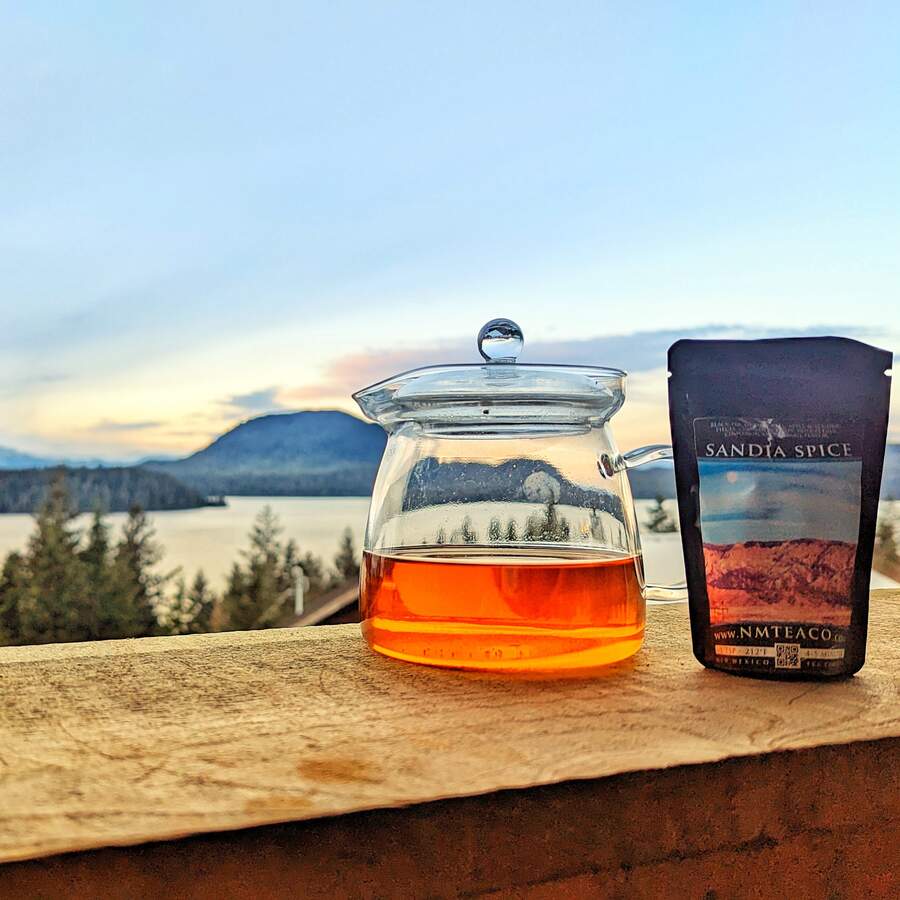 A picture of a glass teapot with Sandia Spice black tea brewed inside it and a bag of the loose leaf tea next to it. In the background is a beautiful landscape with mountains and evergreen trees, taken in Ketchikan, Alaska.