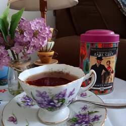 A tea cup and saucer set with purple flowers printed on it and brewed Chocolate Earl Grey tea in it. In the background is a colorful tin of loose leaf tea and a vase with fake purple flowers in it with a tea cup lamp behind it.